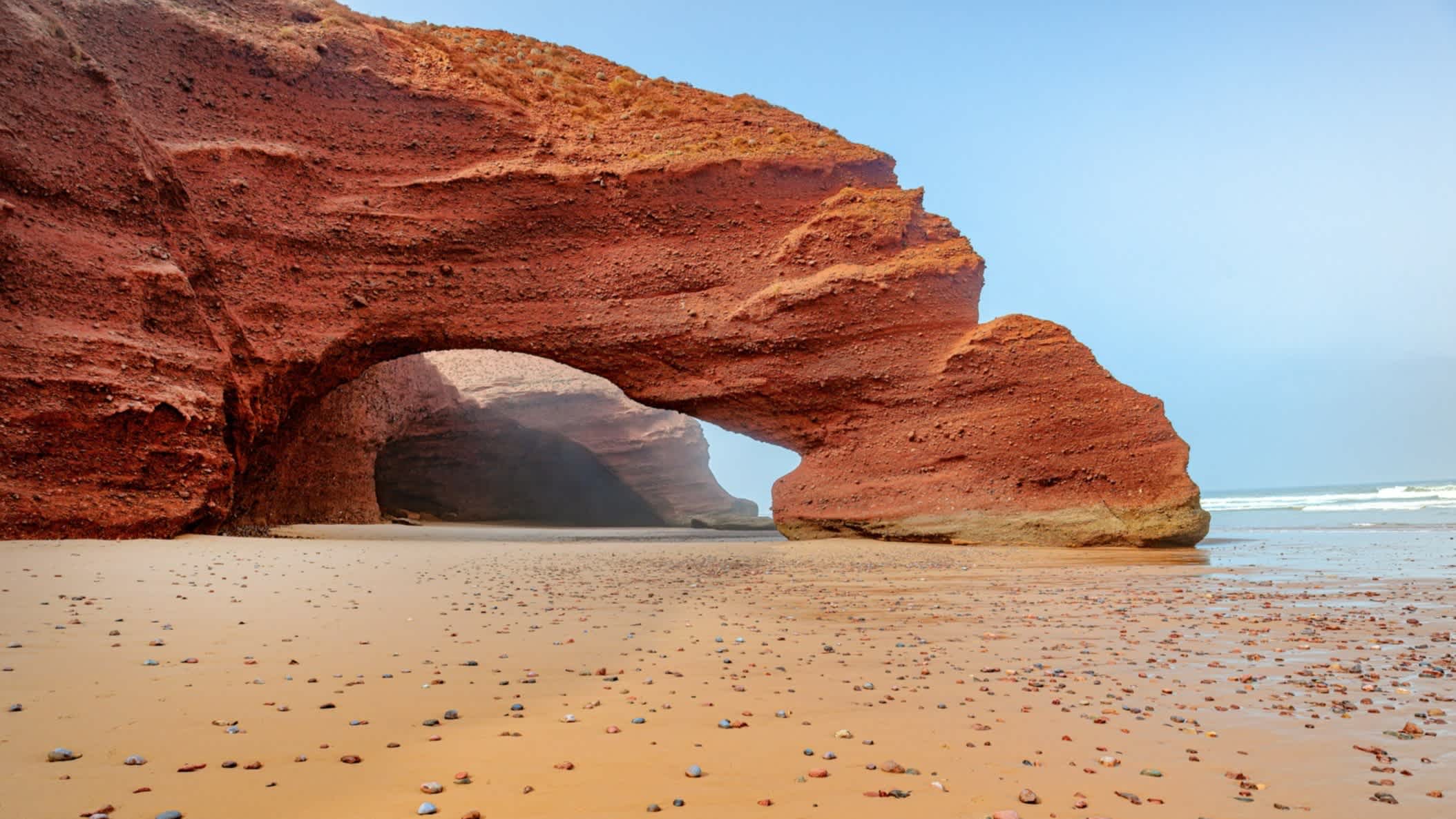 Eine Aufnahme des Strandes von Legzira in Marokko, Nordafrika mit Blick auf einen riesigen roten Steintorbogen und vielen bunten Kieselsteinen am Strand.

