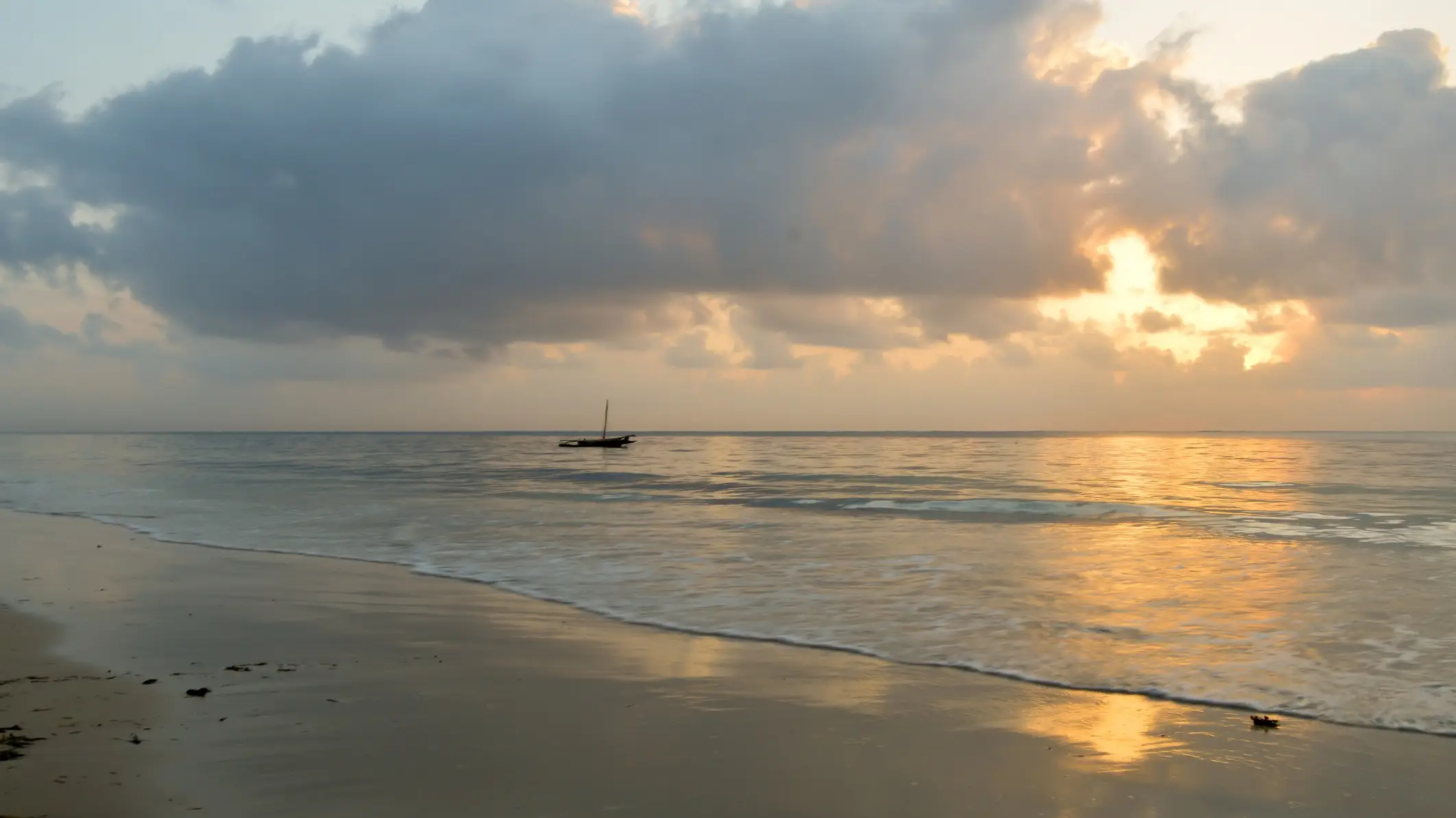 Kenia Bamburi Beach Blick bei Sonnendämmerung auf den Sandstrand Bamburi Beach in Kenia mit einem Segelboot auf dem Wasser und seichten Wellen.