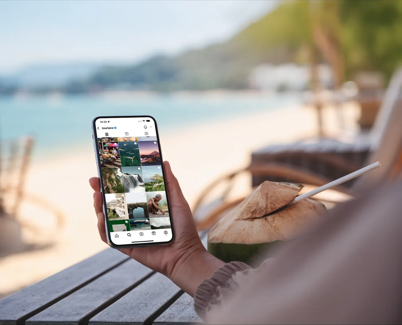Eine Person sitzt an einem Tisch am Strand und hält ein Smartphone in der Hand, auf dem eine Fotogalerie-App angezeigt wird. Auf dem Tisch liegt eine frische Kokosnuss mit einem Strohhalm, und im Hintergrund sind das Meer, der Sand und unscharfe Grünflächen zu sehen.