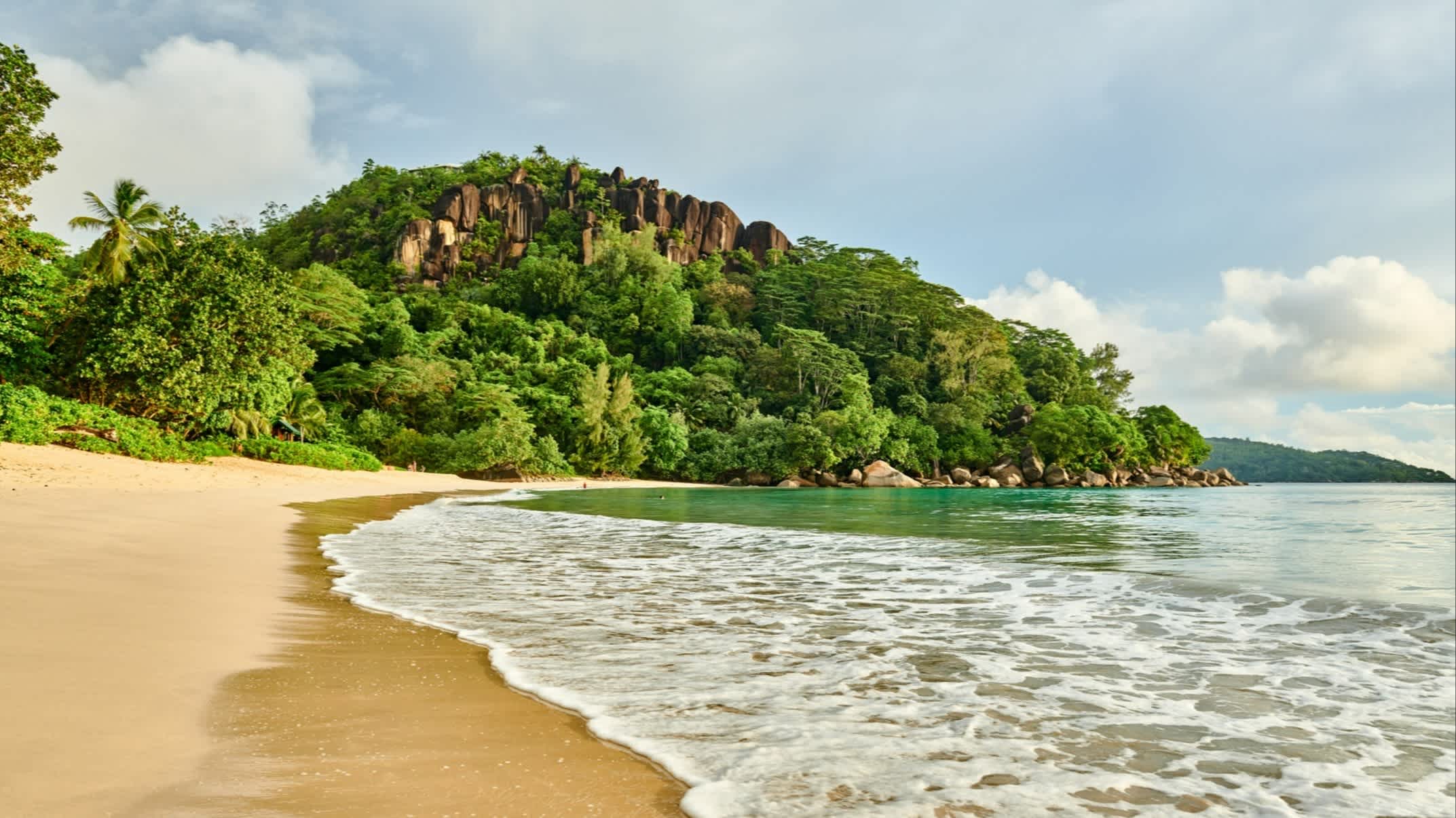 Der schöne Strand Anse Louis auf den Seychellen auf der Insel Mahé mit Blick auf den goldenen Sand, das grüne Meer und die dicht bewachsenen Hügel und Granitfelsen am Rand. 

