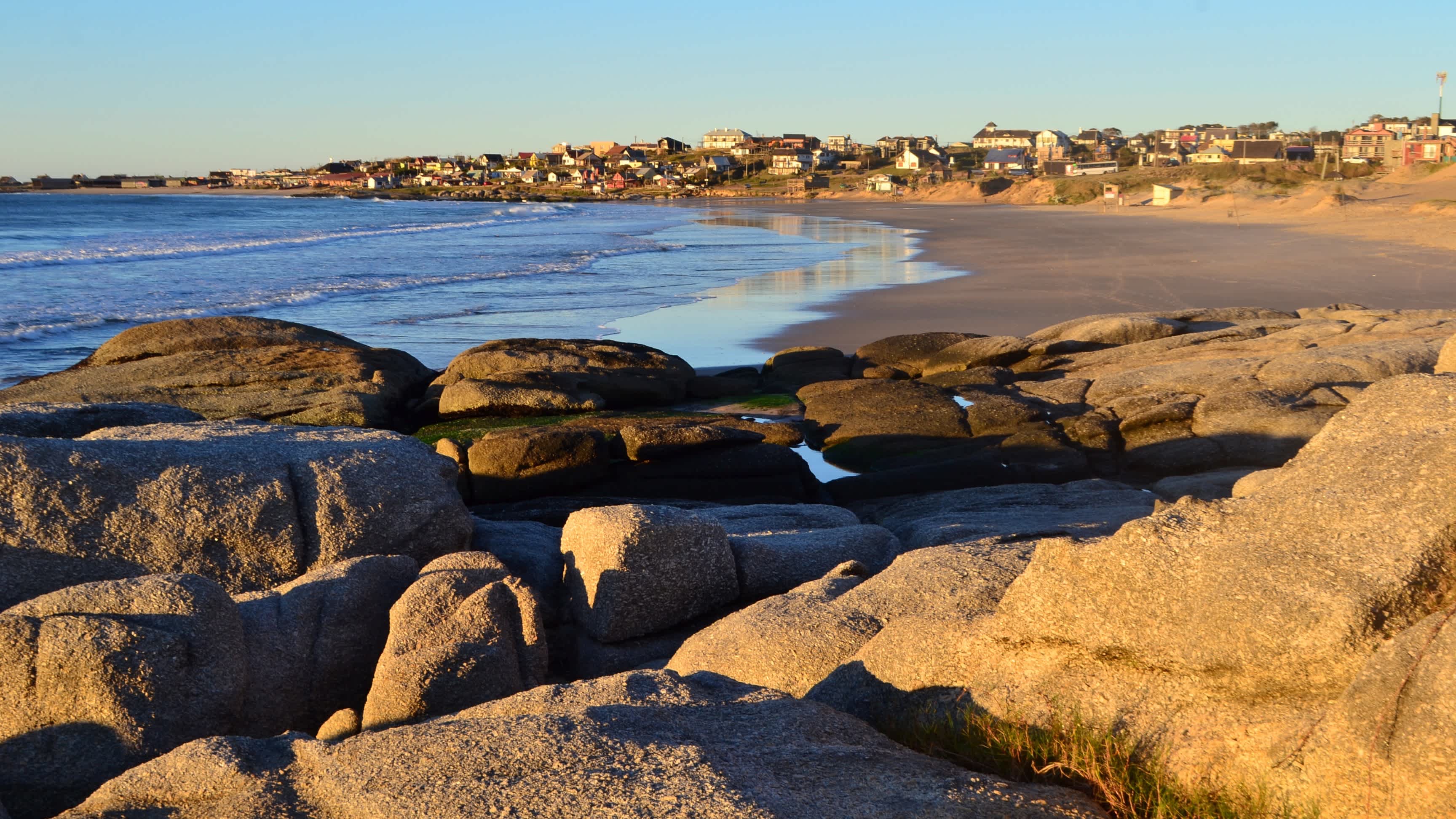Blick auf den Rivero Strand in Punta del Diablo, Uruguay mit Felsen im Bild und bei Sonnenschein sowie die bebaute Küste.