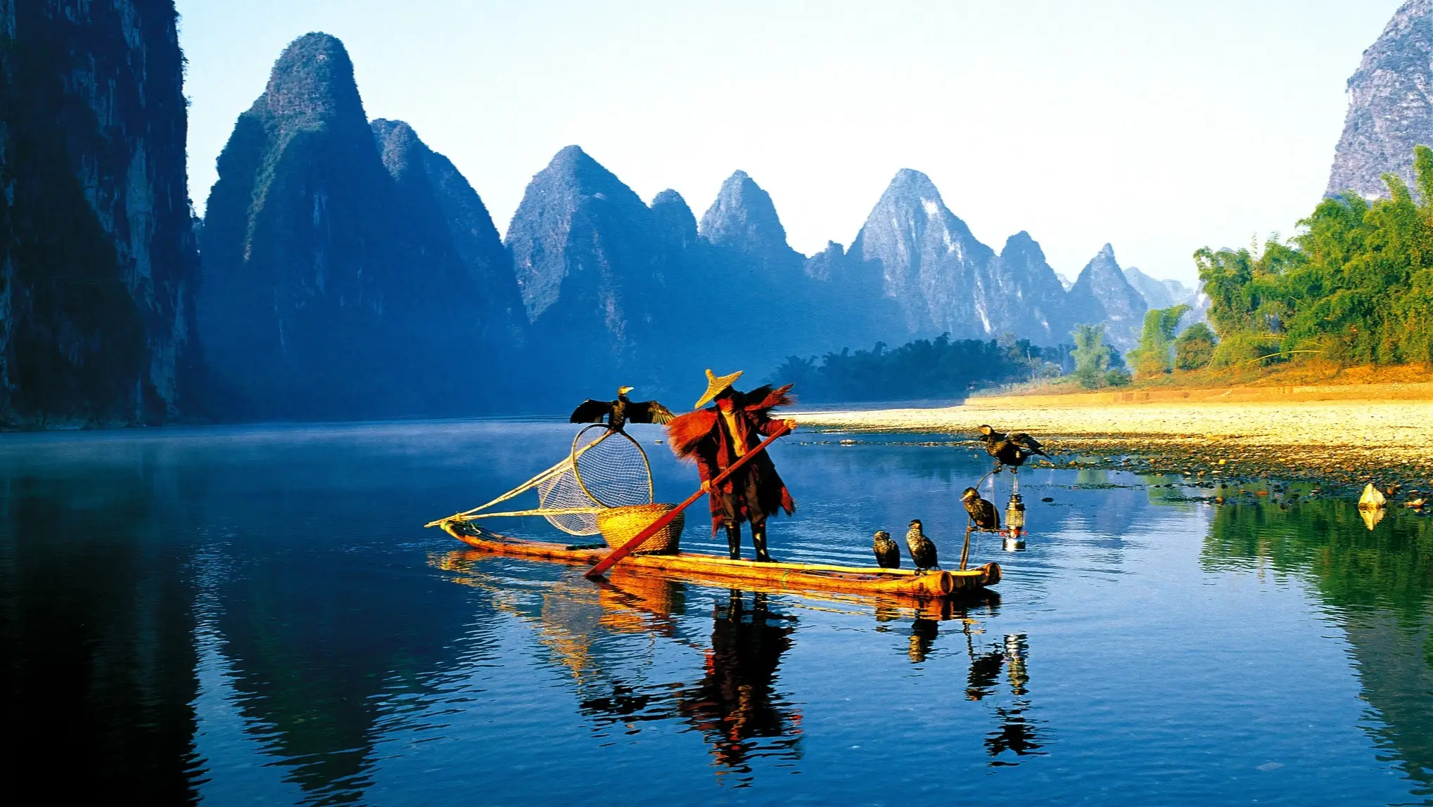 China, Guilin, Fischer Fischer mit Kormoranen auf Bambusfloss in China, umgeben von majestätischen Karstbergen und ruhigem blauen Wasser.