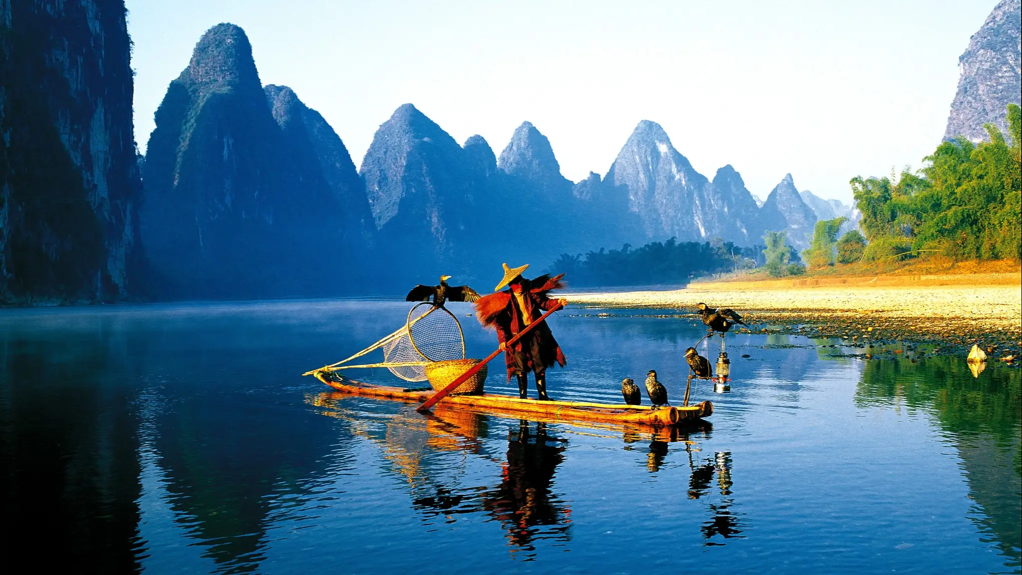 Fischer mit Kormoranen auf Bambusfloss in China, umgeben von majestätischen Karstbergen und ruhigem blauen Wasser.