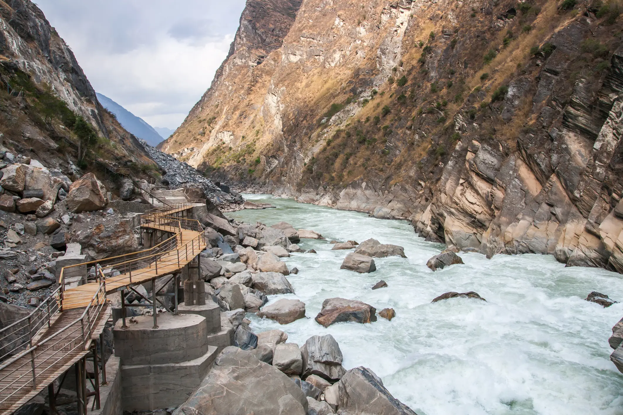 Ein Holzsteg schlängelt sich entlang einer felsigen, engen Schlucht über einem schnell fließenden Fluss, umgeben von steilen, zerklüfteten Bergen unter einem bewölkten Himmel.