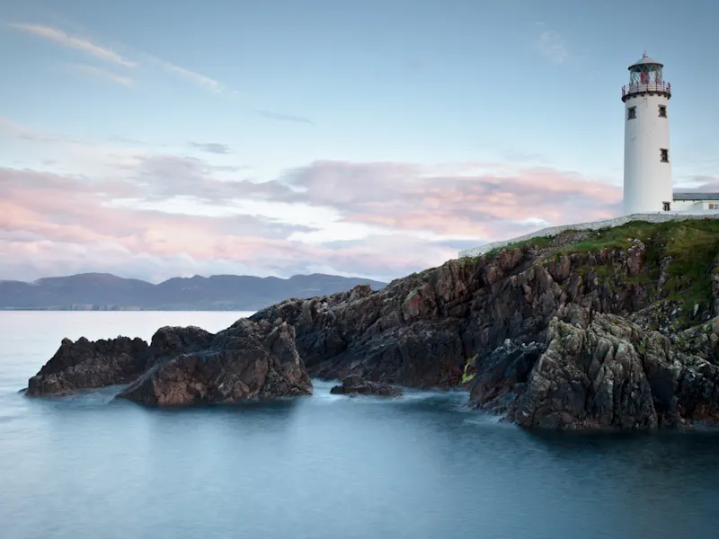 Leuchtturm auf felsiger Küste mit Bergen im Hintergrund. Fanad, Donegal, Irland.