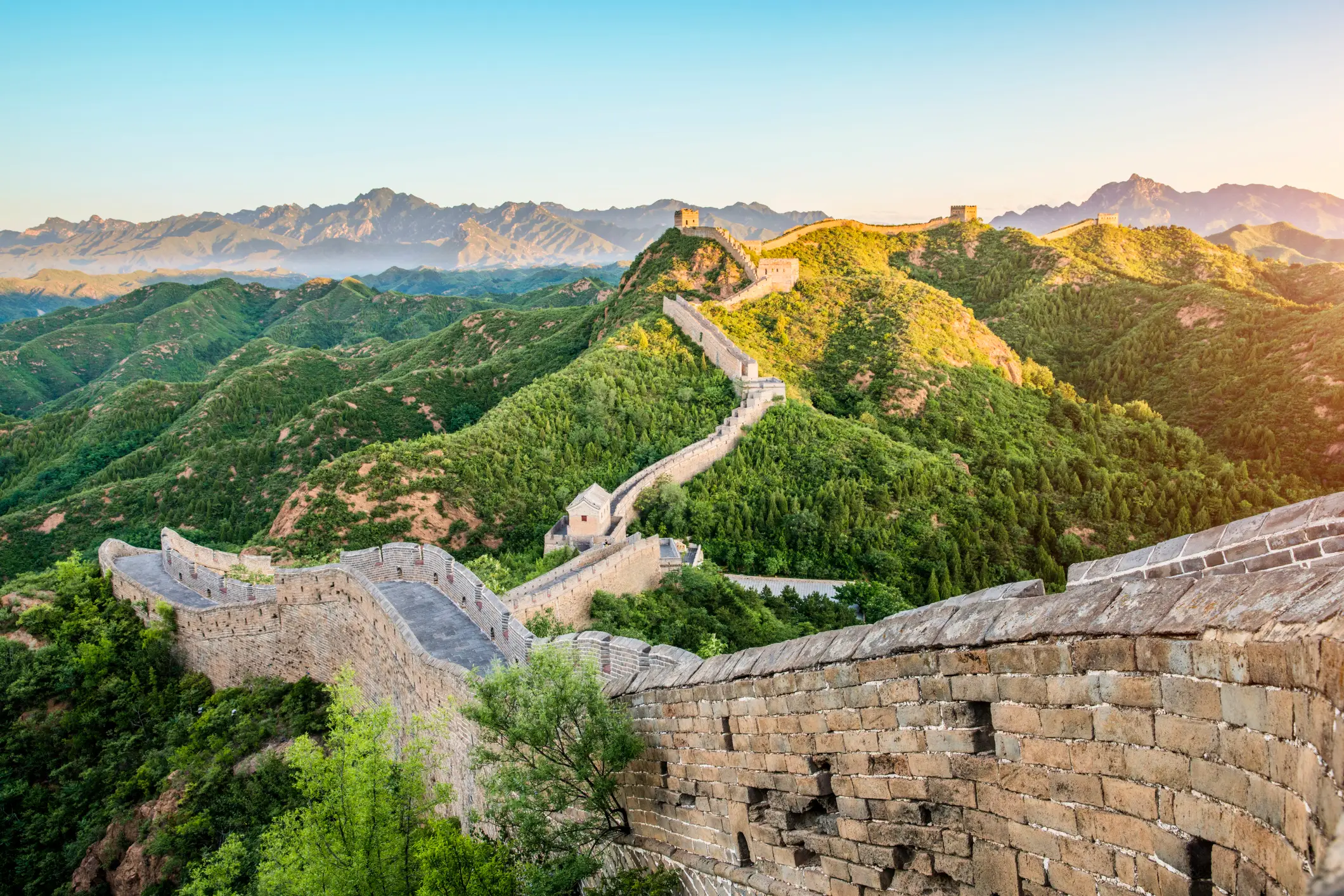 Die Chinesische Mauer schlängelt sich durch grüne Berge bei Sonnenaufgang, Wachtürme säumen den historischen Mauerweg.