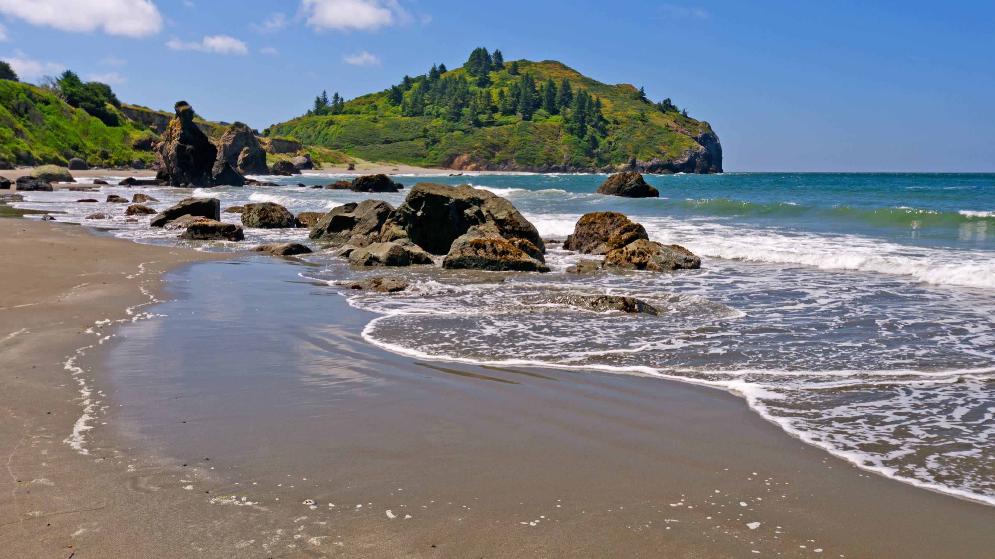 Der natürliche Strand Trinidad State Beach, Humboldt County, Kalifornien, USA bei herrlichem Sonnenschein und mit Blick auf Felsen, das seichte Wasser sowie grünen Hügeln.