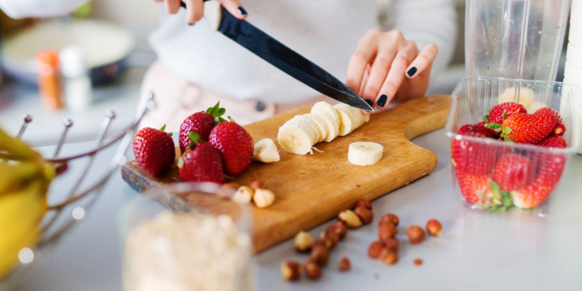 woman cutting a banana on a wooden cutting board next to strawberries, nuts, and a bowl of fruit