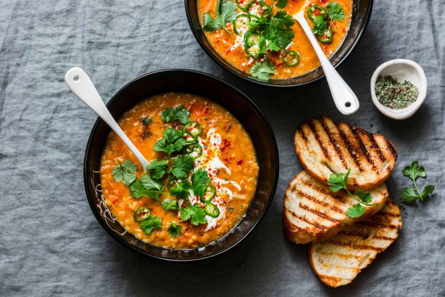 Lentil soup with cilantro in a black bowl with a white spoon next to three pieces of toast on a gray background