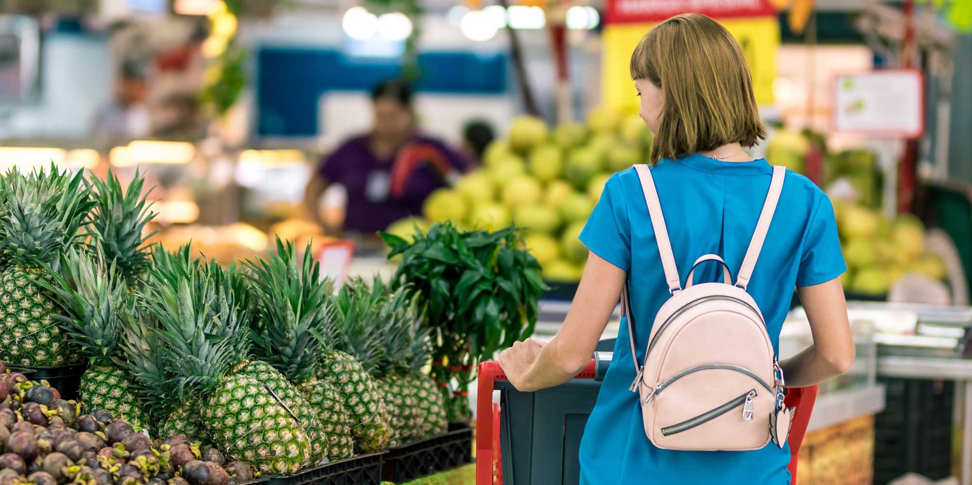 Woman in a blue t-shit and pick backpack pushes her cart around a grocery store next to pineapples.