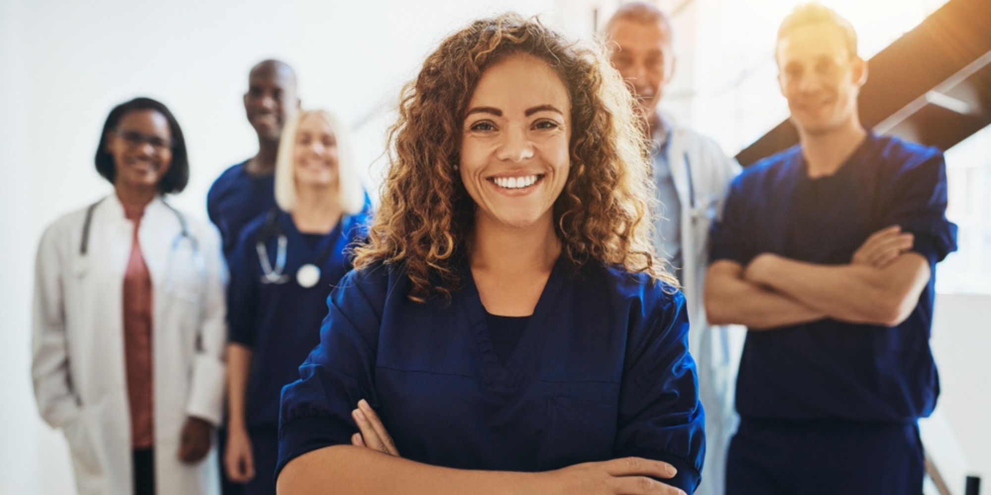 Young female doctor in navy scrubs smiling with arms crossed, other medical professionals lined up in a row behind her.