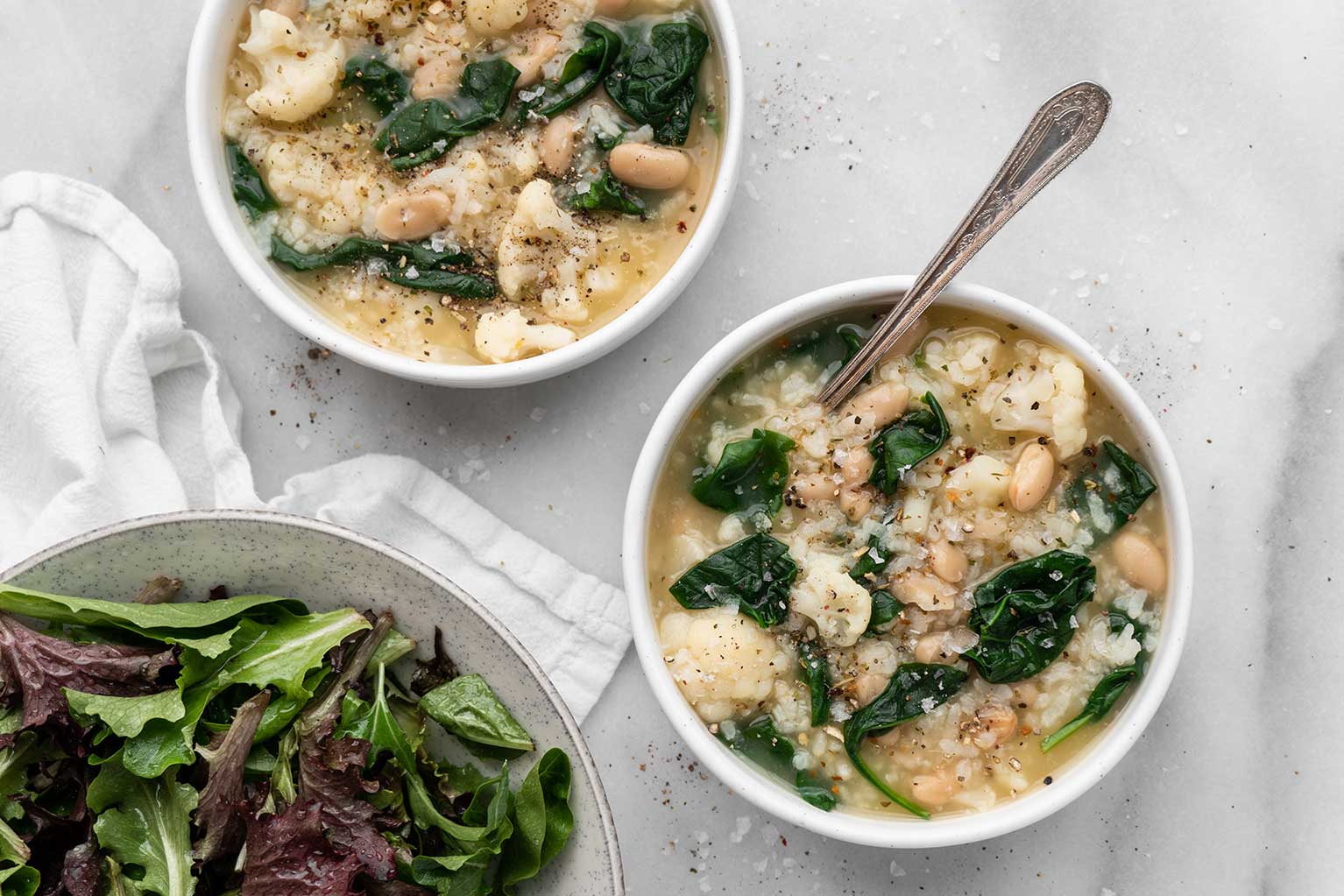 Two bowls of rice and white bean soup with spinach and side salad on counter