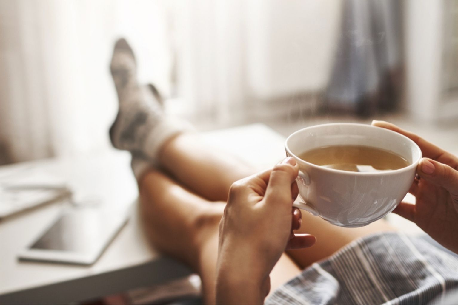 Person holding a cup of tea while sitting on the couch with their legs up on the coffee table.