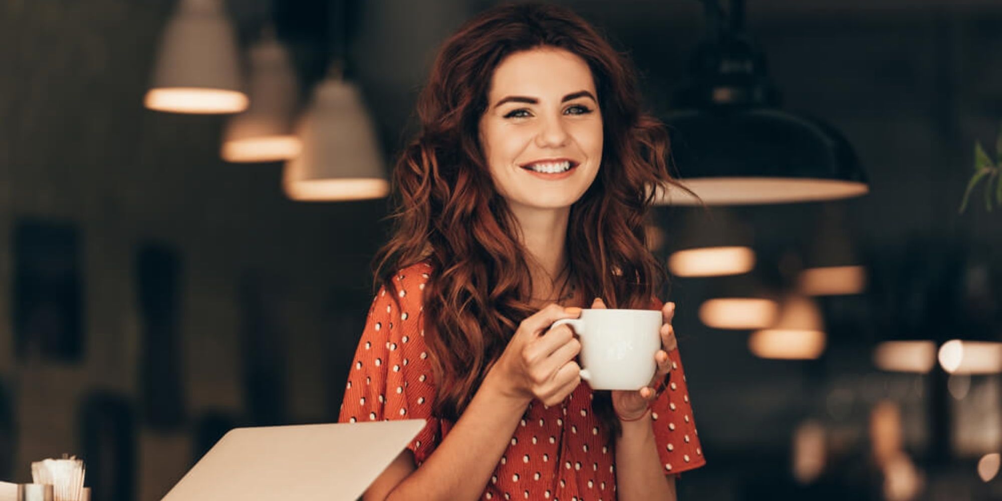 Woman at coffee shop holding a white mug next to a laptop.