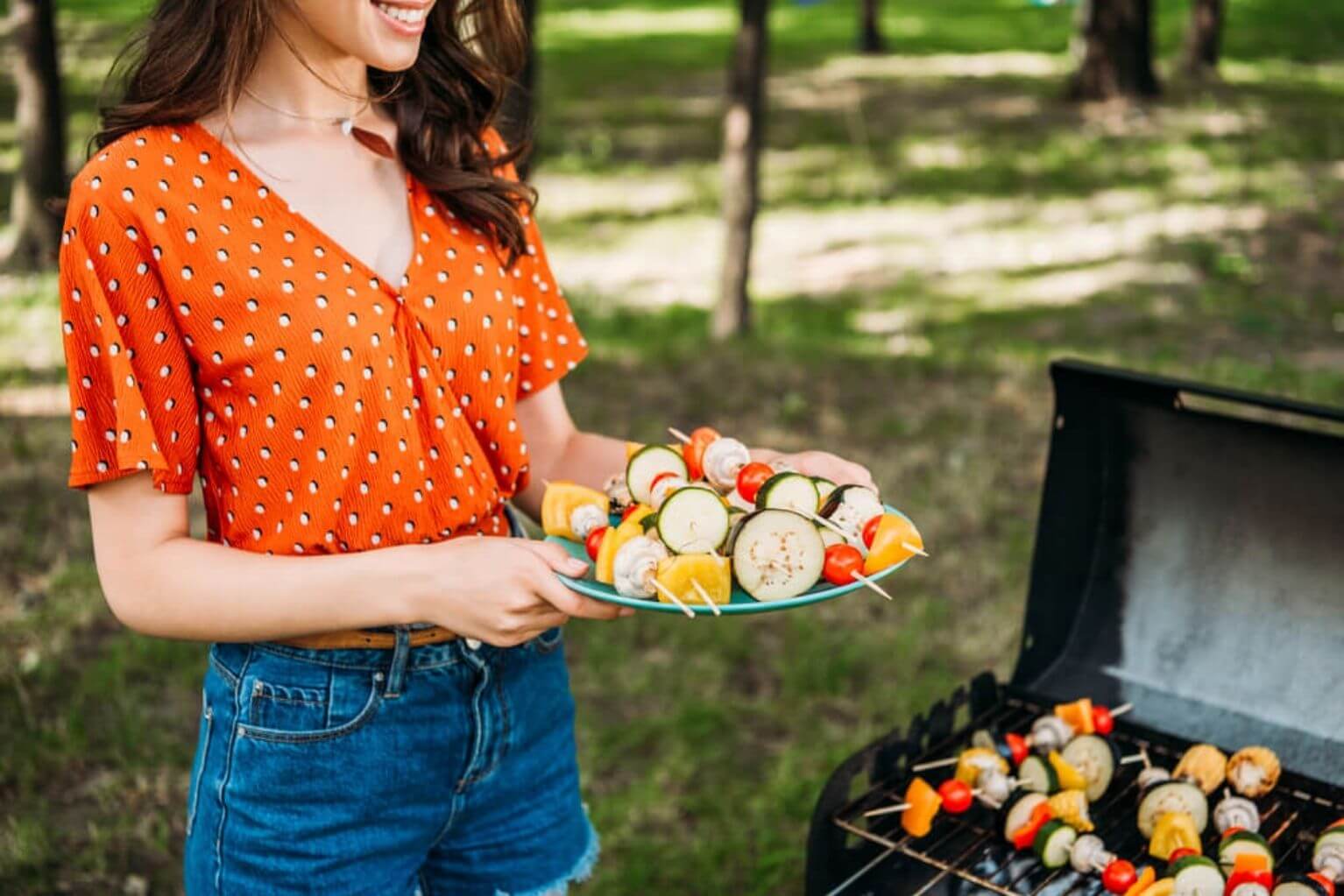 Woman in an orange shirt holding vegetable skewers next to a grill outside. 