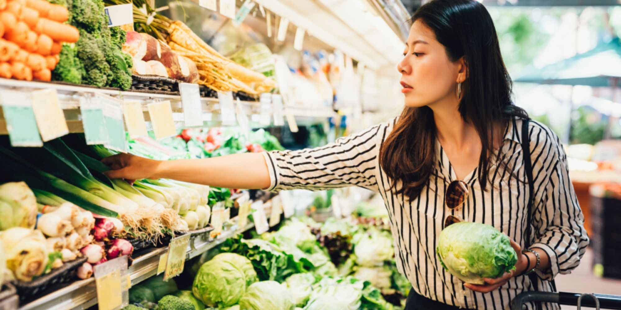 woman shopping at the grocery store holding a head of cabbage and reaching for green onions