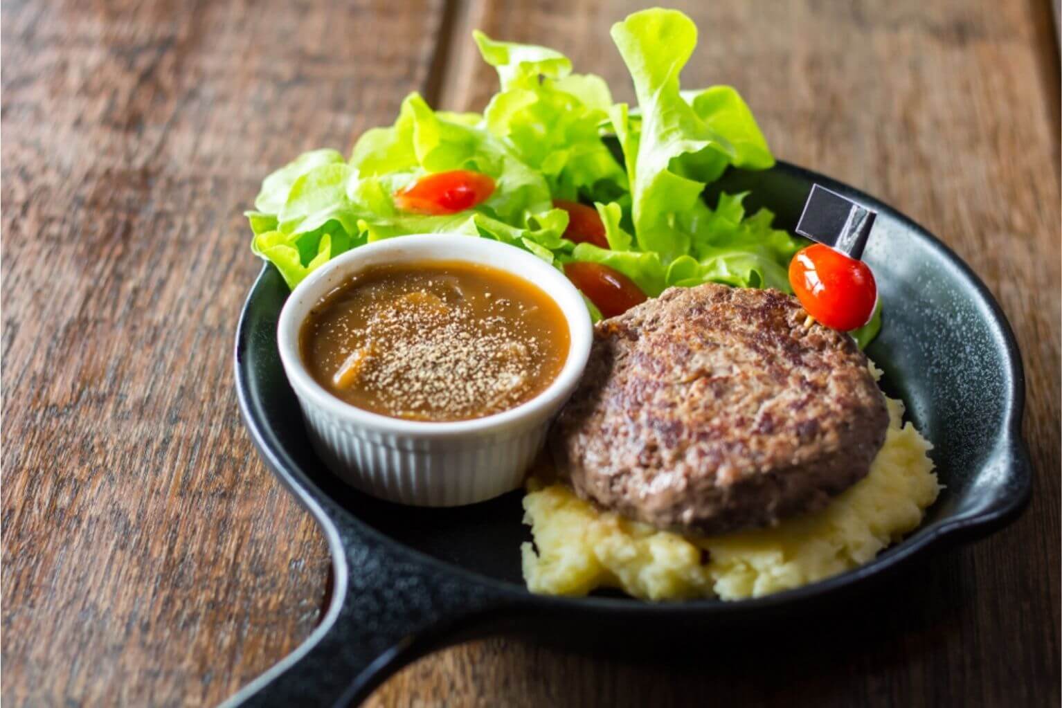 Cooking skillet filled with mashed potatoes, a beef patty, a side salad and a white cup full of low carb gravy, all sitting on a wooden table.