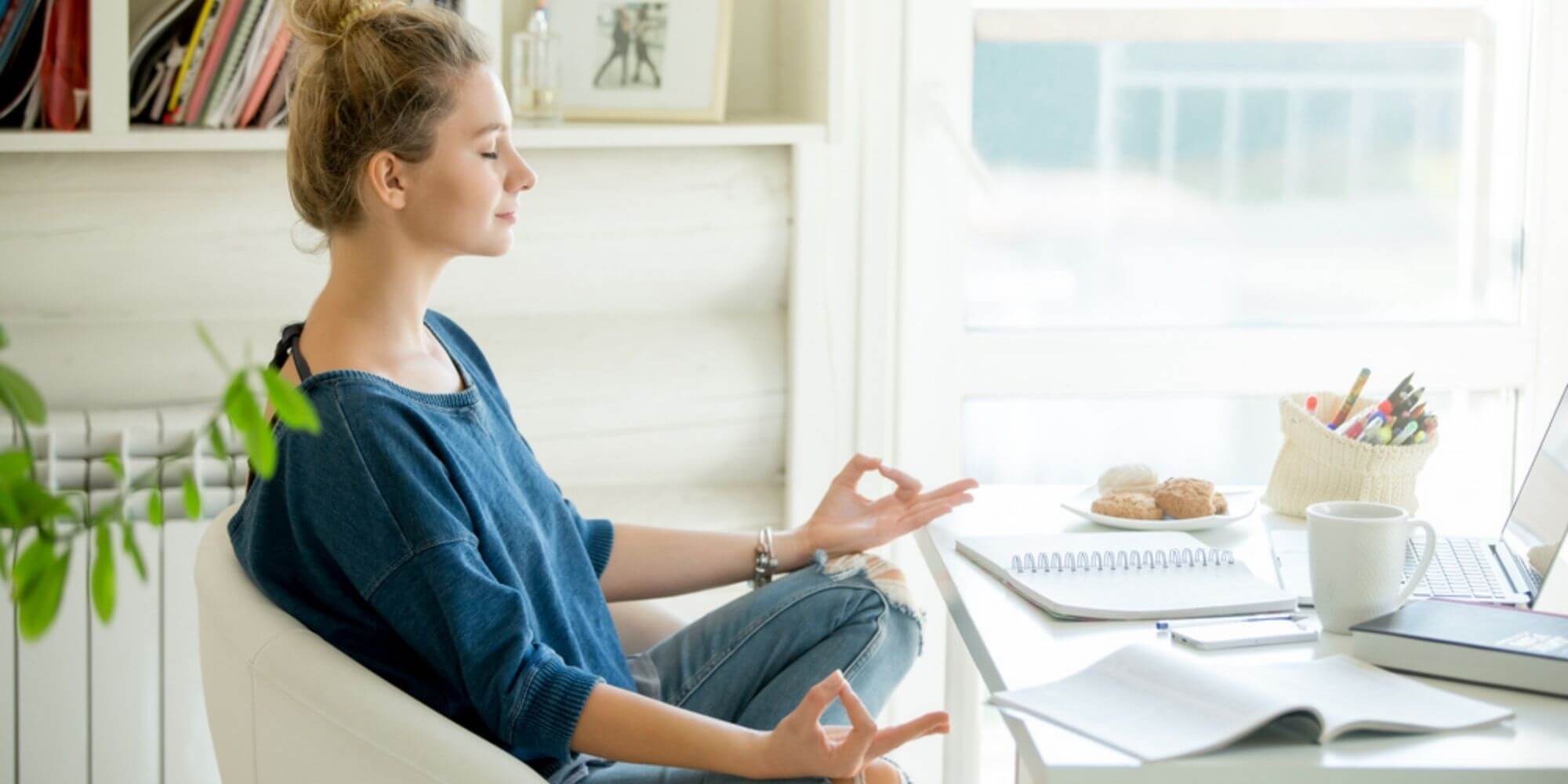 Woman meditating on white chair next to desk with a computer, coffee cup, and work papers.