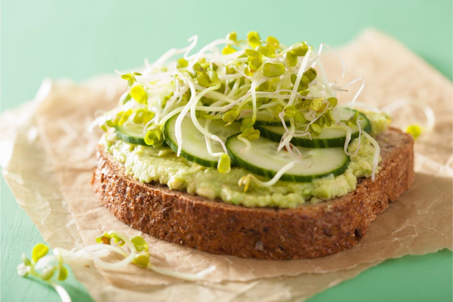 One slice of whole wheat bread with avocado, cucumber and sprouts on a piece of parchment paper.