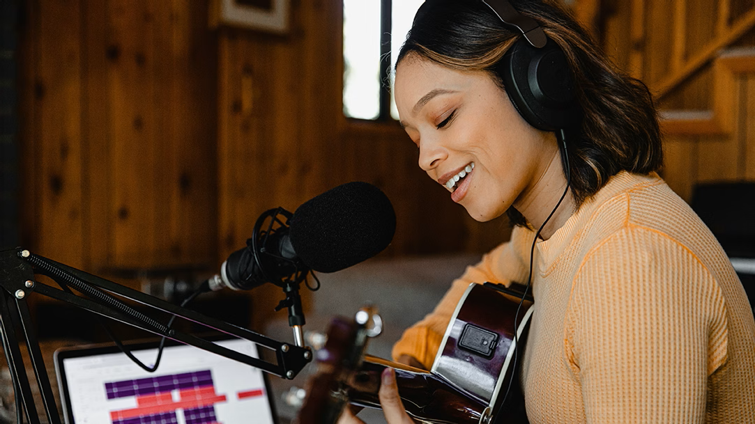 A woman wearing headphones sings into a microphone while strumming a guitar in front of a laptop