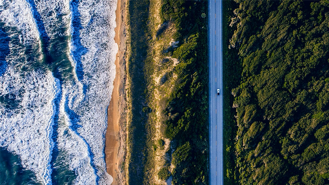 A top-down view of a car on the road between the ocean and a forest along the coast