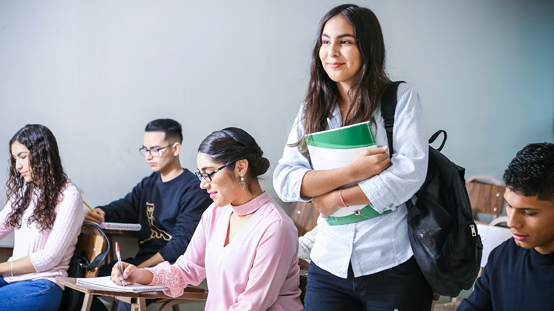 A college student carrying a green and white textbook and wearing a black backpack finds her seat in a classroom filled with other students writing in notebooks
