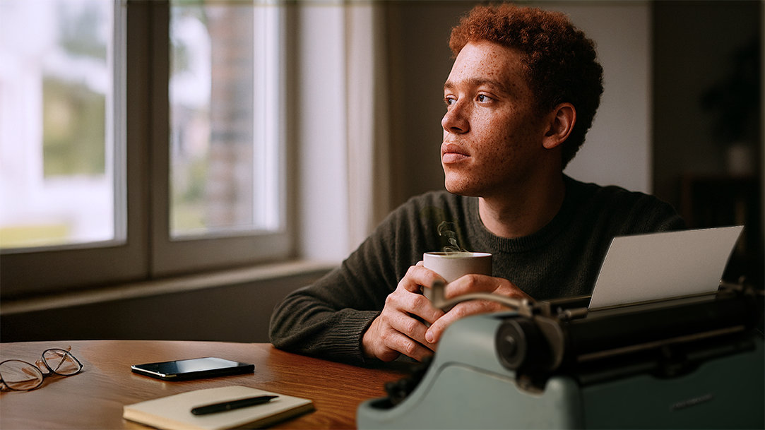 A young man struggling with writer’s block while sitting holding a cup of coffee at a desk that has a pair of glasses, his cellphone, a notebook, and vintage typewriter