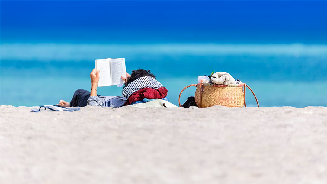 A person reads a book on the beach while lying down on a towel with a woven beach bag next to them