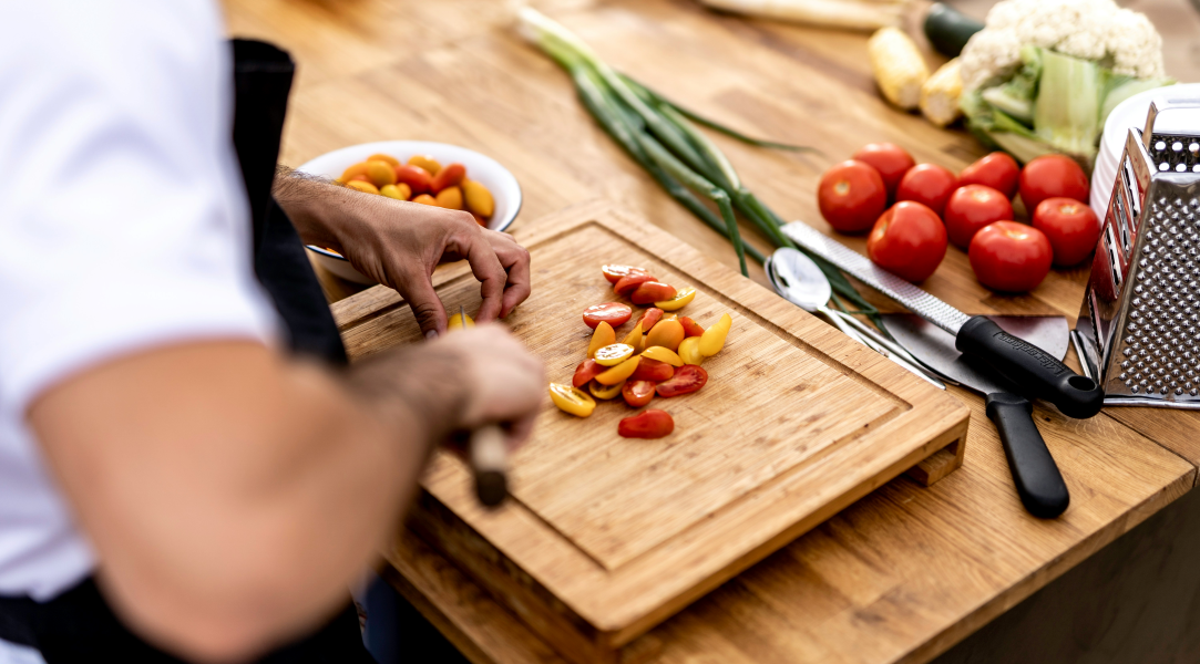 A person cutting up cherry tomatoes on a cutting board with other vegetables on the table