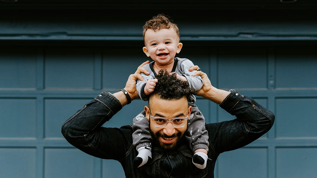 A smiling dad with his smiling baby on his shoulders in front of a dark teal background