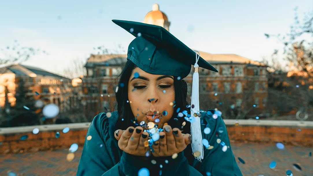 A college graduate wearing a green cap and gown and blowing confetti into the camera