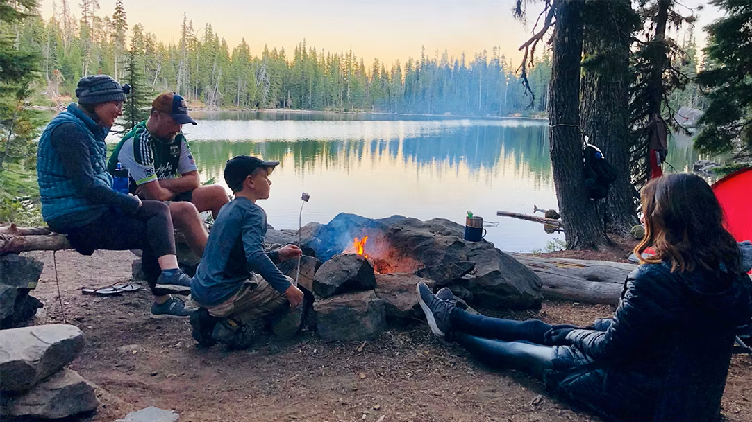 An image of a family at a campfire in the forest next to a lake