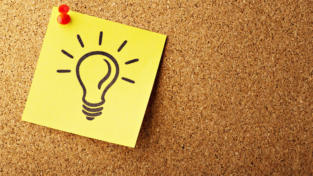 A sticky note with a pen illustration of a lightbulb is pinned to a corkboard with a red pin