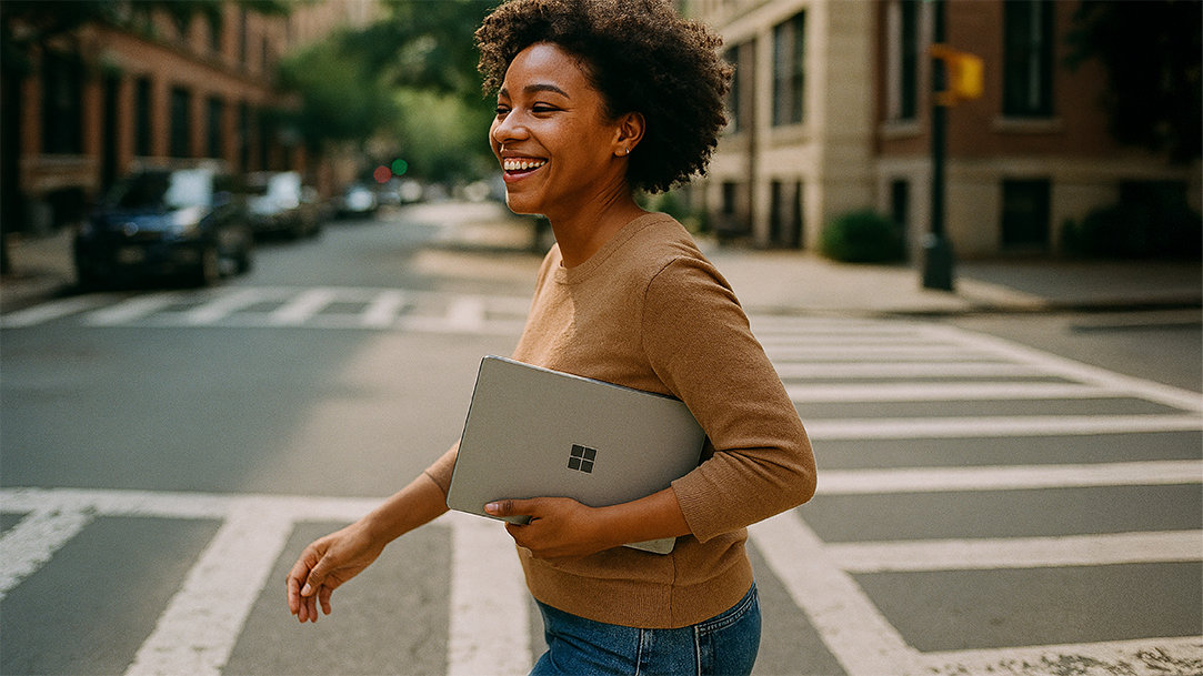 A happy woman walking across a street while carrying a Windows Surface laptop under her arm