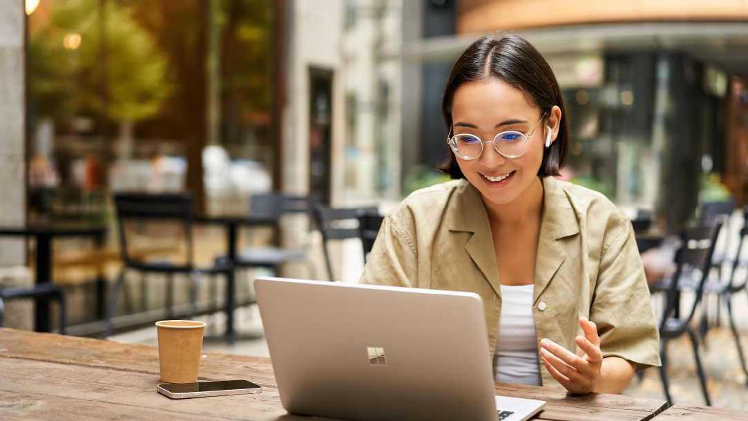 A person wearing glasses smiles and works on a laptop at an outdoor cafe
