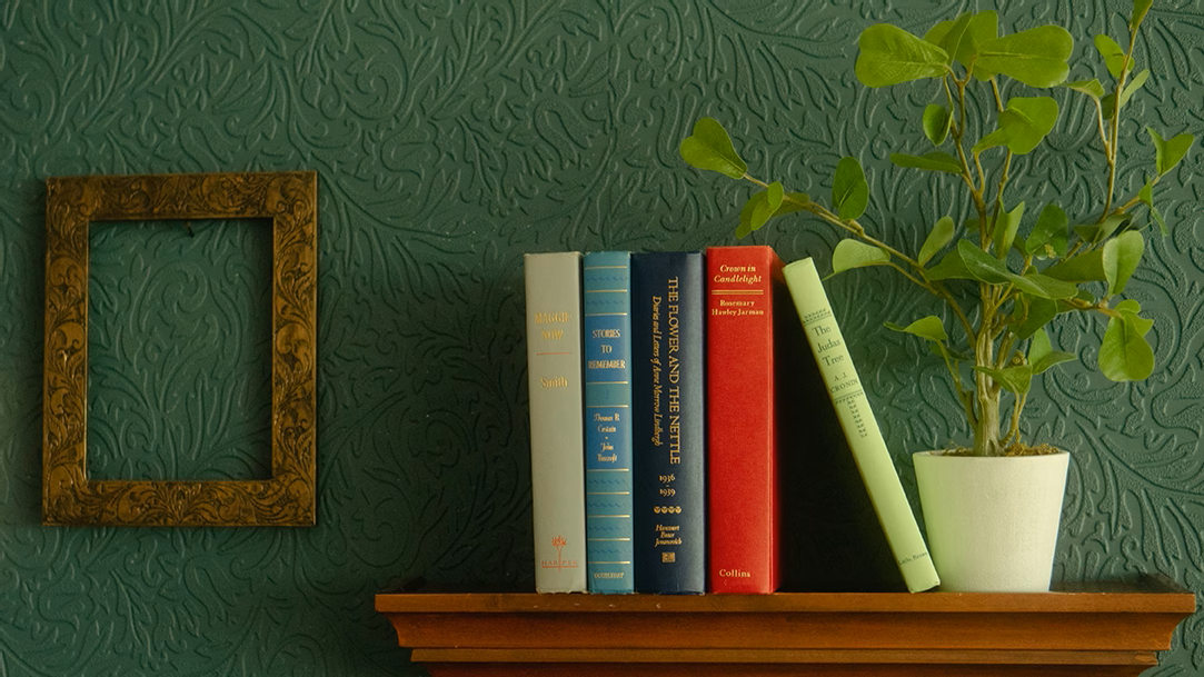 Five books on a brown bookshelf next to a potted plant and an empty picture frame on a green wall