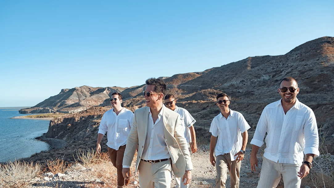 Five men smile while walking on a rocky trail above the ocean during the day