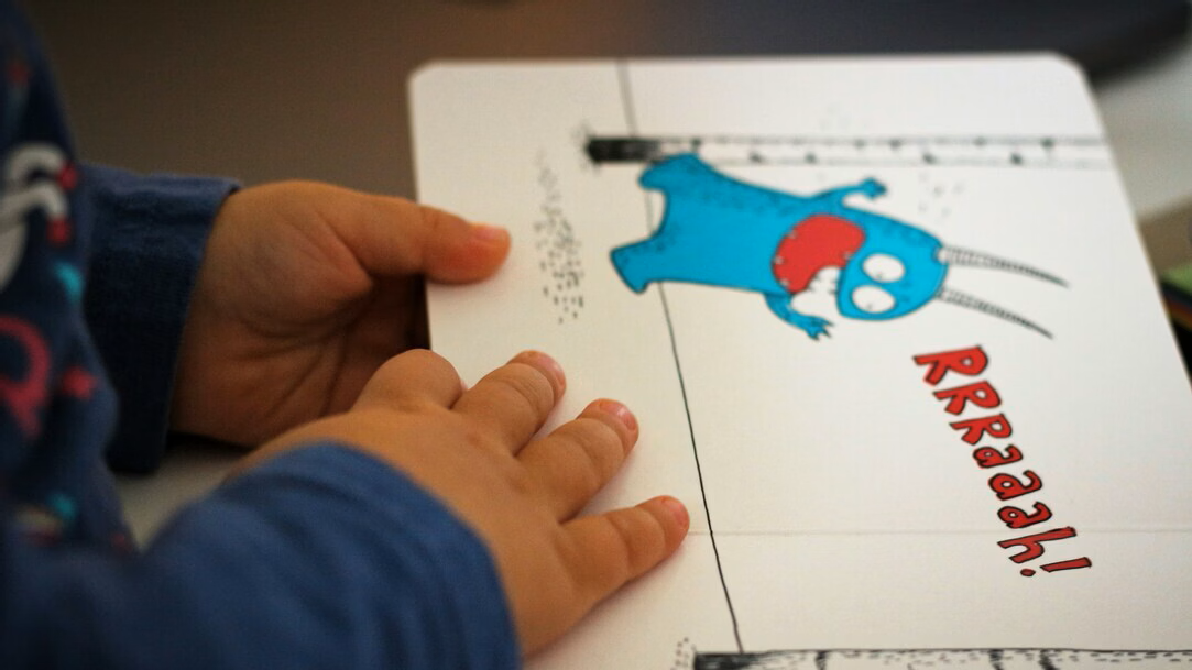 A child’s hand rests on the page of a picture book with an image of a blue monster on a white background