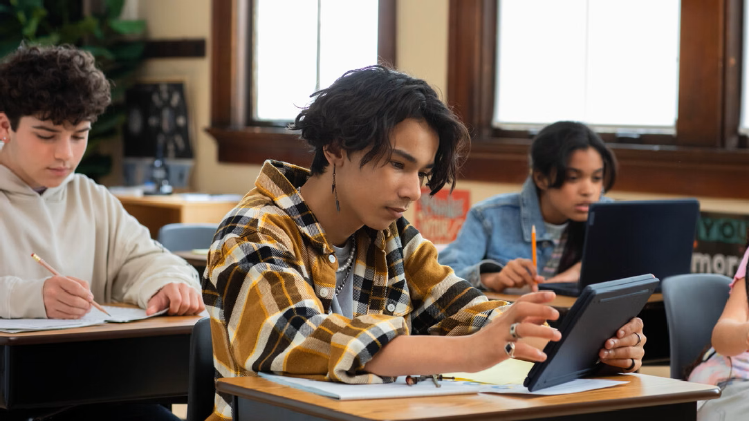 A male student sits at a desk with a laptop. Other students work in the background on laptops and worksheets