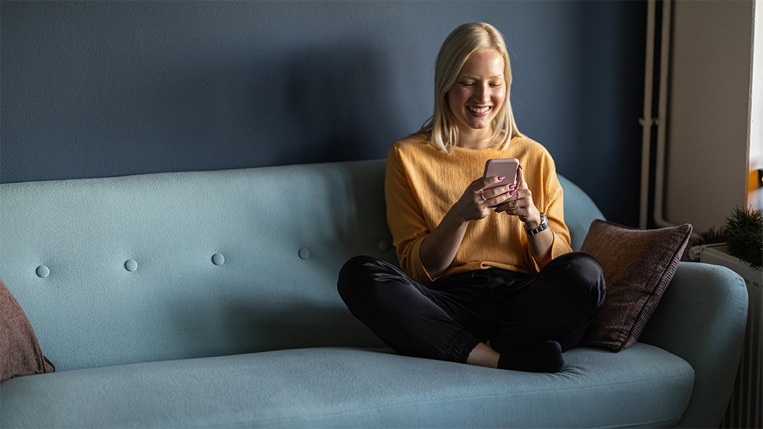 A young woman uses her smartphone to chat with her AI companion while sitting on a blue couch in her living room