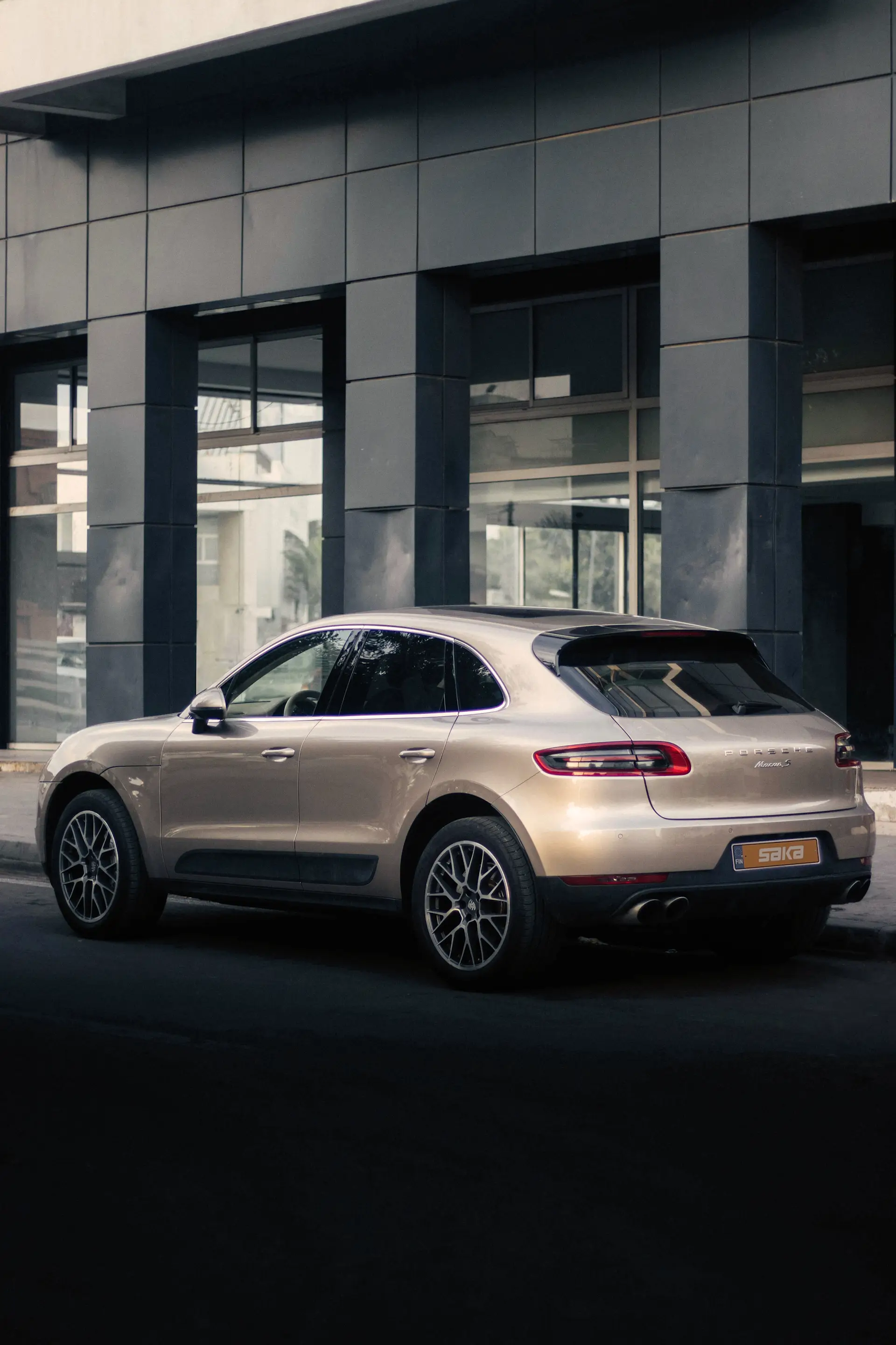 Samppanjanvärinen Porsche Macan -katumaasturi pysäköitynä modernin rakennuksen edustalle hämärässä valaistuksessa.