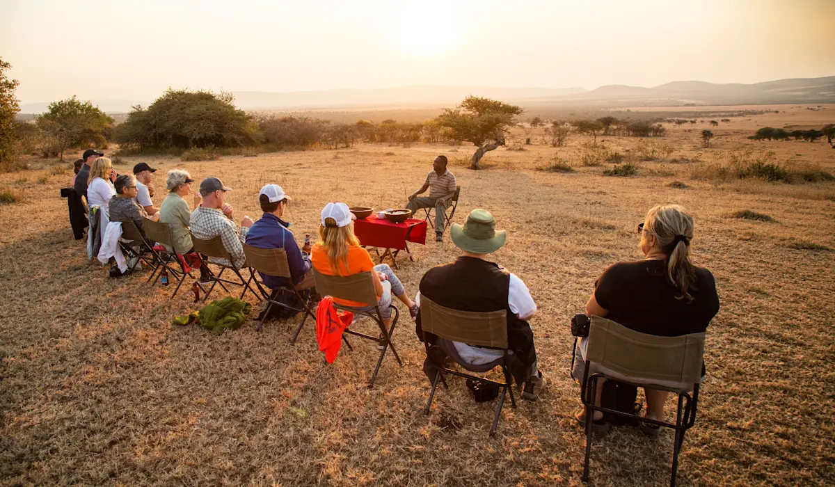 Travelers seated in a semicircle on the plains at sunset listening to a Maasai storyteller
