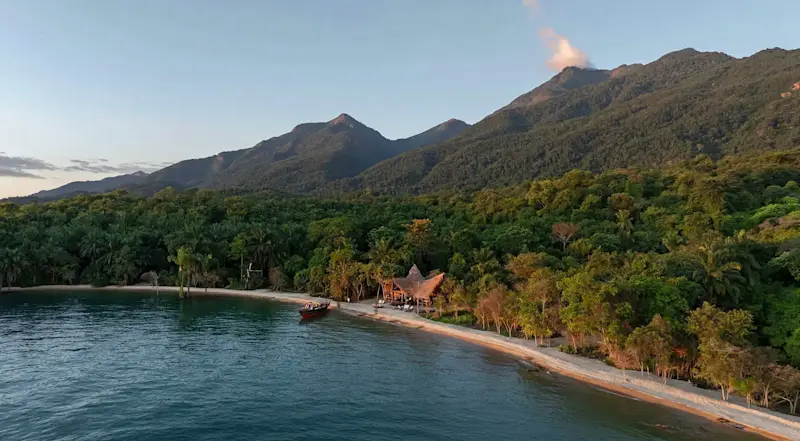 Aerial view of a forested lakeshore beneath the Mahale Mountains, with a lodge and boat on the beach