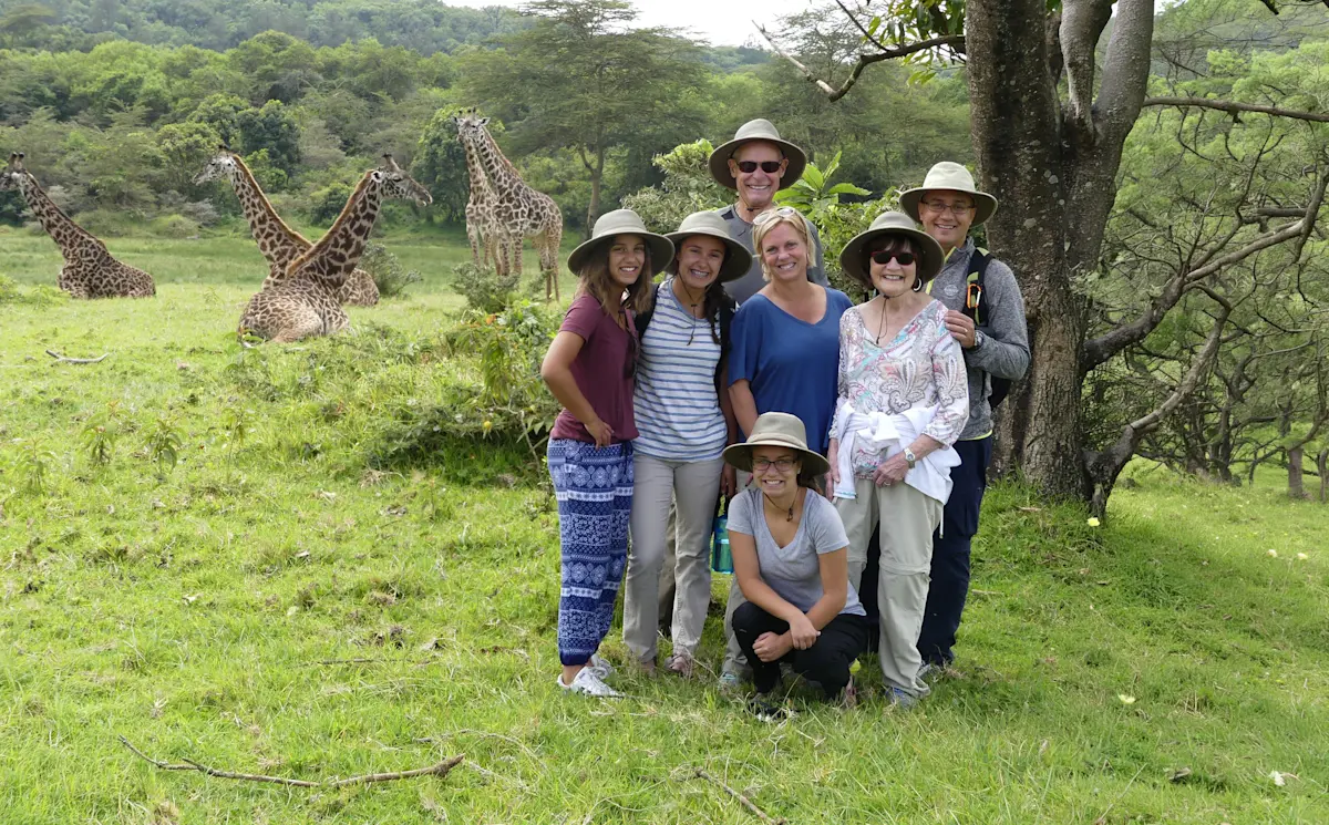Group of travelers in safari hats posing on the grass with giraffes behind them on a walking safari
