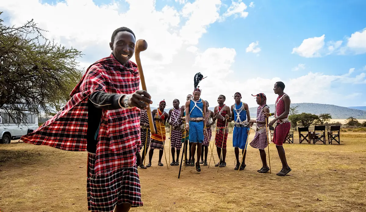 Smiling Maasai man in a red shuka with Maasai warriors dancing behind him in the Eastern Serengeti