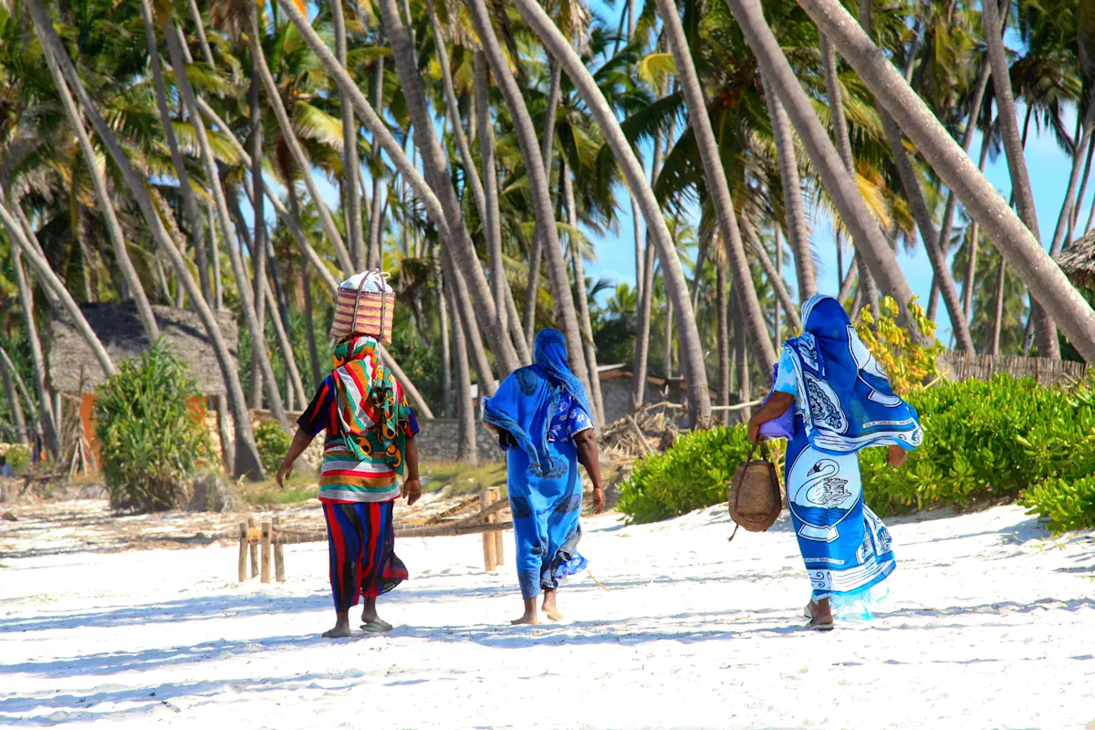 Women in colorful wraps walking along a white sand beach under palm trees in Zanzibar