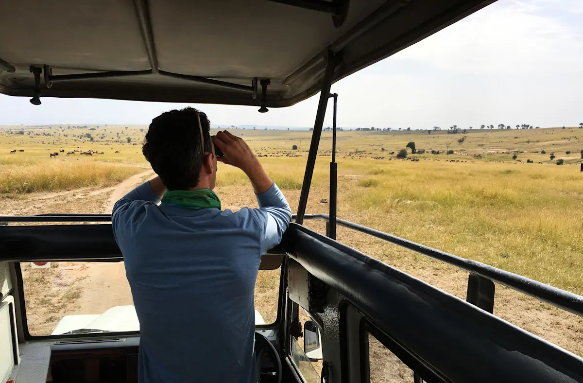 Guest standing in an open-roof safari vehicle looking through binoculars over the plains