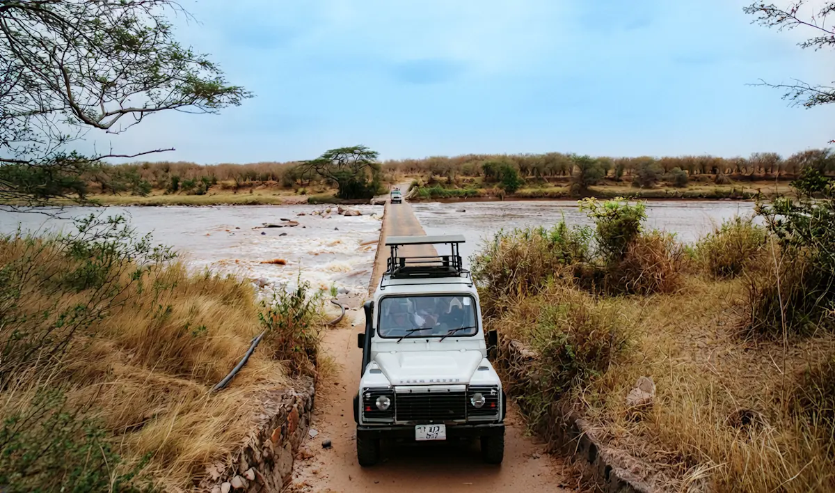 Safari vehicle crossing a narrow causeway over a river, seen from behind