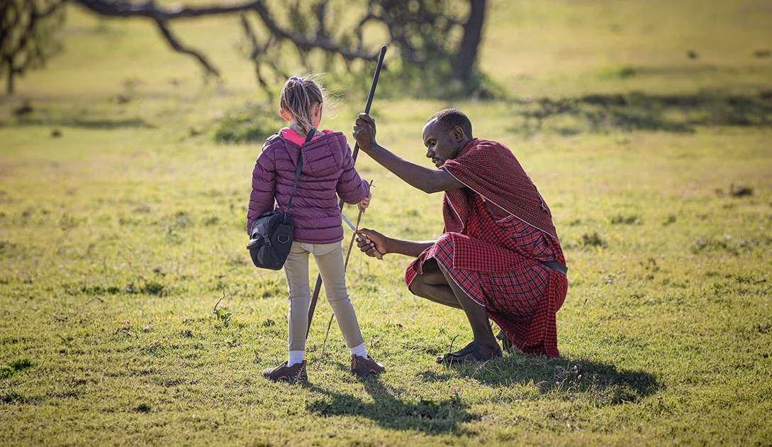 Maasai man kneeling to show a young girl how to hold a spear