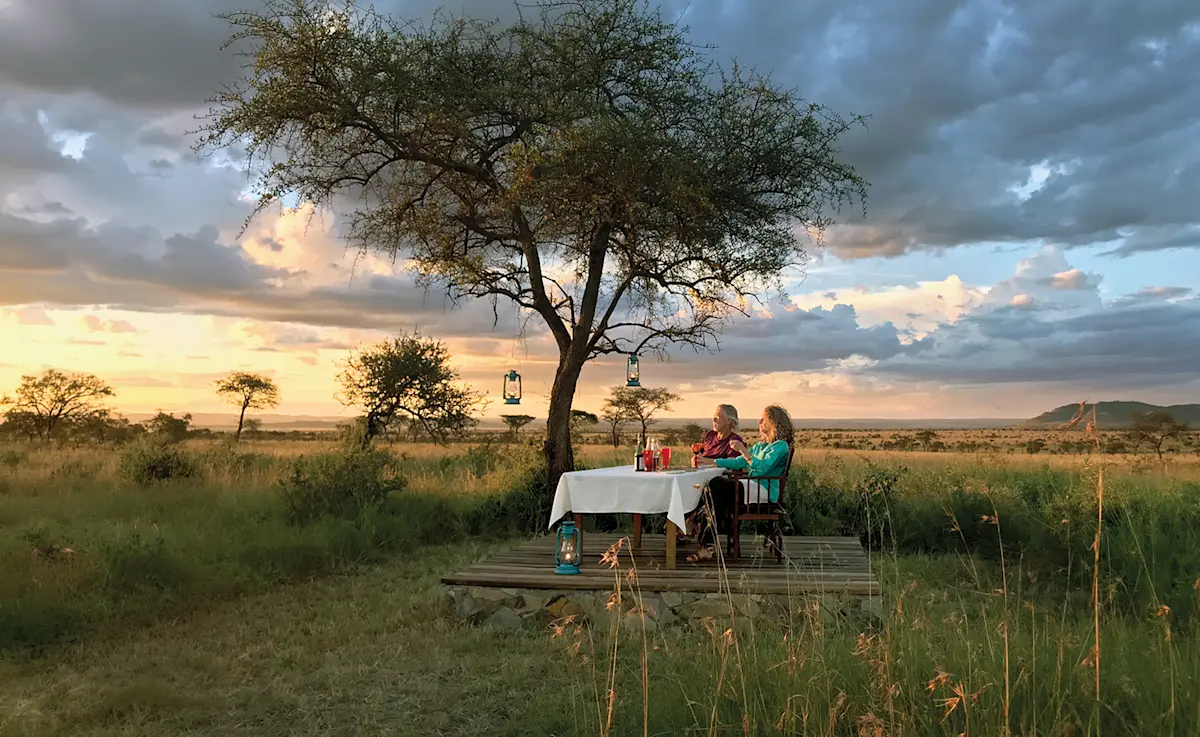 Rick Thomson and Judi Wineland enjoying sundowners at a table under an acacia tree, Serengeti Nyumba